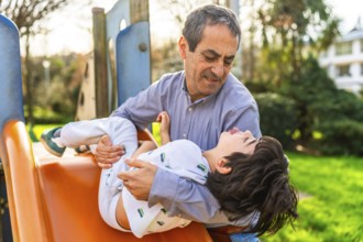 Grandfather catching his grandson sliding down the slide at a playground, having fun together on a