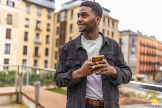 African smiling young man sending a message using smartphone standing int the city