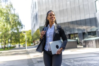 Confident latin businesswoman walking out the office carrying laptop along the city