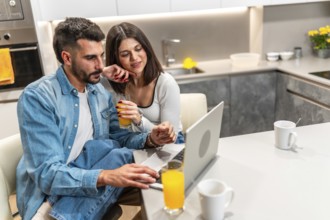 Young couple sitting at kitchen table, drinking orange juice and using laptop, enjoying their time