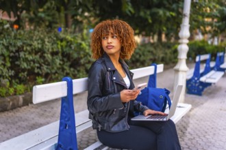 Latin young female student using smartphone and laptop sitting on park bench