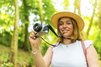 A young woman holding camera in forest smiling at camera among green trees