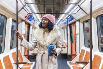 Young black woman wearing winter coat and pink woolen hat holding handrail and reusable coffee cup