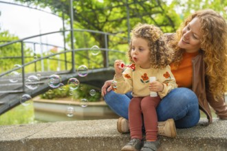 Blonde adorable playful girl blowing bubbles in the park with her mother
