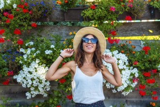 A woman wearing a straw hat and sunglasses is standing in front of a wall of flowers. She is