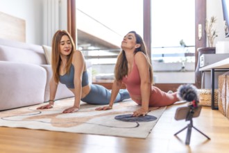 Two women practicing bhujangasana or cobra pose during online yoga class at home living room