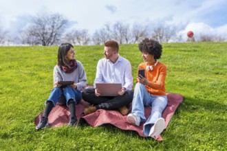 Three university students using electronic devices while sitting on a blanket in a park, enjoying a