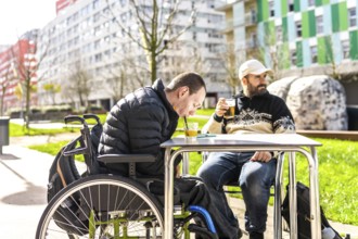 Two men enjoy drinks at an outdoor cafe, one using a wheelchair, demonstrating inclusivity and