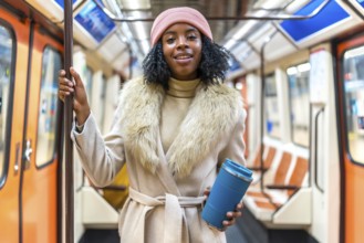Smiling young black woman wearing winter coat and pink wool hat holding reusable coffee cup while