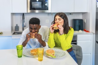 Young multiethnic couple eating homemade burgers in their modern kitchen, enjoying a casual meal