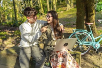 Two students are working on a laptop in a park, charging it with a power bank while sitting on a