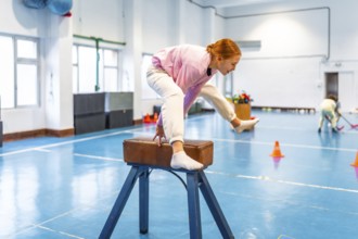 Girl practicing gymnastics, balancing on pommel horse in school gym, showing determination and