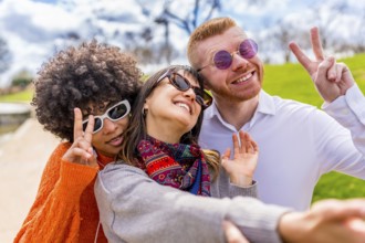 Three happy multi ethnic friends wearing sunglasses gesturing peace sign while taking a selfie in a