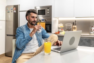 Man savoring a cup of coffee while working remotely on a laptop in a stylish, modern kitchen,