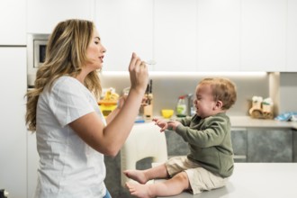 Young mother struggling to feed her crying baby boy while sitting on the kitchen counter at home,