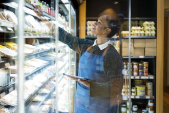 Smiling saleswoman wearing blue apron checking food products using digital tablet while working in