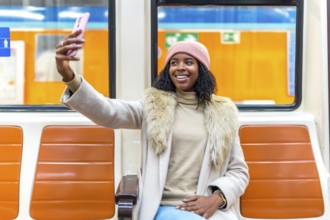 Smiling young woman wearing winter clothes taking a selfie while traveling on subway train