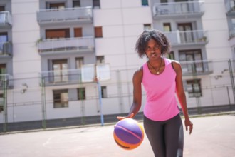 Confident afro woman looking at camera while dribbling in an urban basketball court