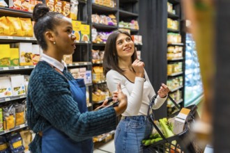 Saleswoman using tablet, advising customer holding shopping basket, choosing products on shelves in