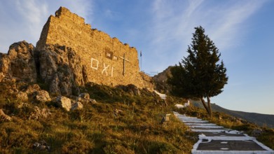 Warm evening light, stone wall of a ruin next to a tree under a blue sky with OXI lettering,