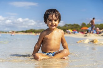 A young boy is sitting in the water with his arms crossed. He is smiling and he is enjoying himself