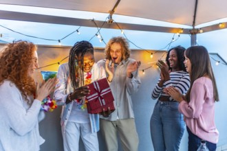 Happy young african man opening birthday gift with multi-ethnic friends during roof party