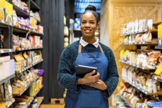 Confident saleswoman in an apron, smiling while holding a digital tablet and working among shelves