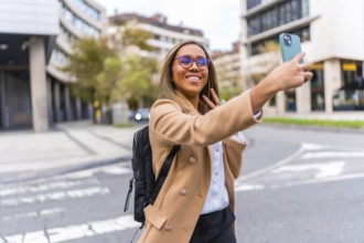 Happy latin businesswoman taking selfie and gesturing success using smartphone standing in the city