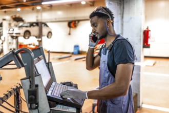 Young mechanic using a laptop and talking on the phone while working on a car in a professional