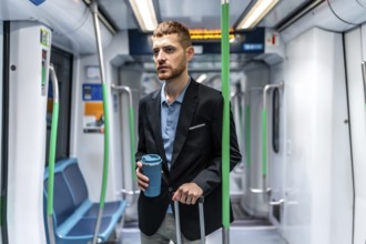 Young businessman commuting to work by subway, holding a reusable coffee cup and a trolley bag