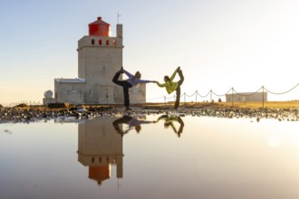 Two people are doing yoga on a beach in front of a lighthouse. The water is calm and the sky is
