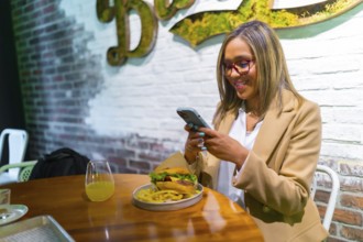 Beautiful latin woman wearing business casual clothes enjoying tasty burger and texting on the