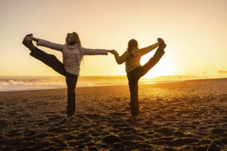 Two women are doing yoga on a beach. The sun is setting in the background, creating a warm and