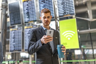 Businessman using smartphone while holding reusable coffee cup in airport, checking flight schedule