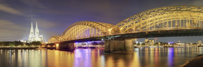 Panorama of the illuminated Cologne Cathedral with Hohenzoller Bridge at night, with reflection of