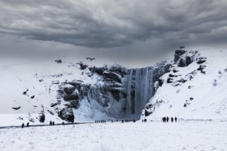 Tourists at Skogafoss waterfall, snowy and icy rock face, dark clouds above, Sudurland, Iceland,