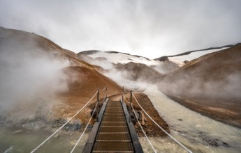 Bridge over steaming stream between colourful rhyolite mountains with snowfields, Hveradalir