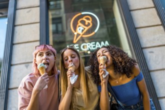 Low angle view photo of three diverse female friends enjoying eating a delicious ice cream outside