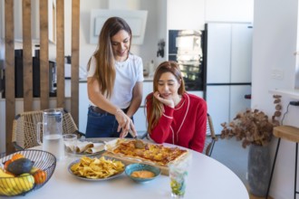 Two women are slicing and sharing a rectangular pizza, nachos with sauce and tacos at home in their