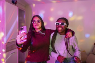Two happy teenage girls with sunglasses taking a selfie with smartphone at a home party with disco