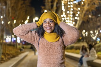 Smiling young woman wearing winter clothes adjusting her yellow wool cap under christmas lights in