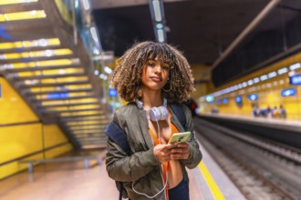 Latin young woman waiting in the platform of underground metro using phone and carrying headphones