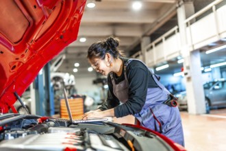 Young woman mechanic using a laptop while examining a car engine in a professional workshop