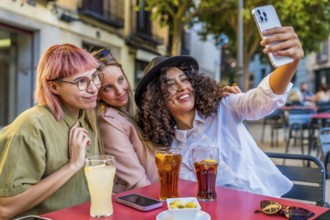 Happy female friends taking selfie together sitting on a sidewalk cafeteria drinking soda and