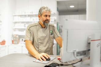 Mature male pharmacist in uniform using computer in the store