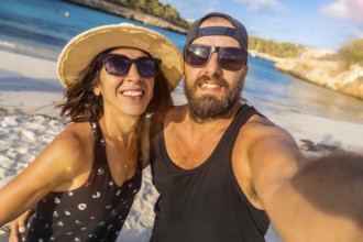 A man and woman are smiling for the camera on a beach. The man is wearing a black tank top and