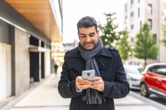Elegant man in winter clothes sending message and smiling walking along the city