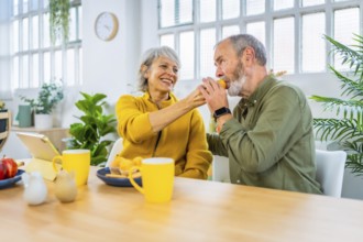 Senior woman feeding her husband in a romantic gesture in the morning sharing breakfast at home