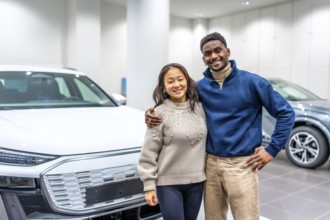 Happy multiethnic couple hugging, buying new electric car at dealership, embracing modern eco