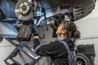 Skilled female mechanic working under a lifted car, inspecting and repairing the brake system in a
